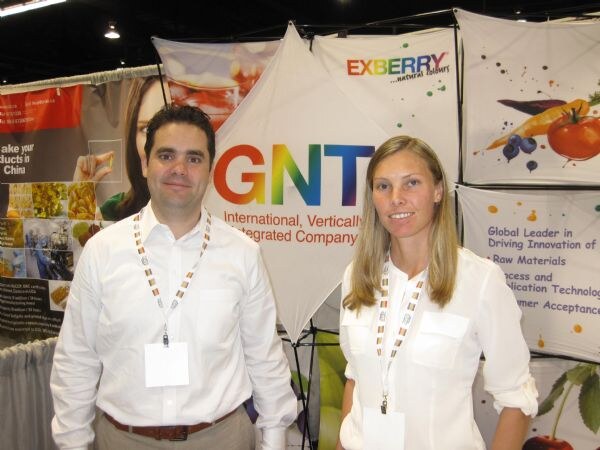 A man in a white shirt and a woman with long blonde hair, both wearing badges, stand in front of a booth with a large GNT sign and an Exberry banner.