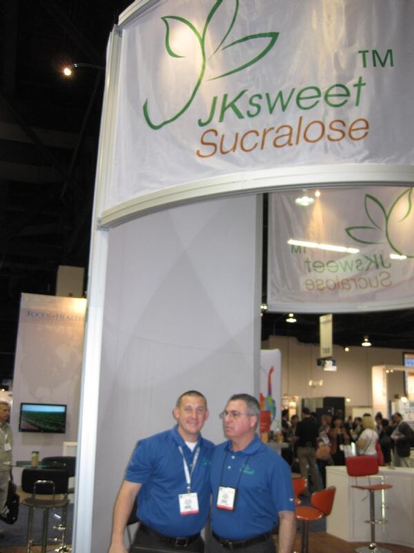 Two men in blue polo shirts and badges stand beneath a white JKsweet Sucralose banner with a green leaf logo, with other exhibit booths and attendees behind them.