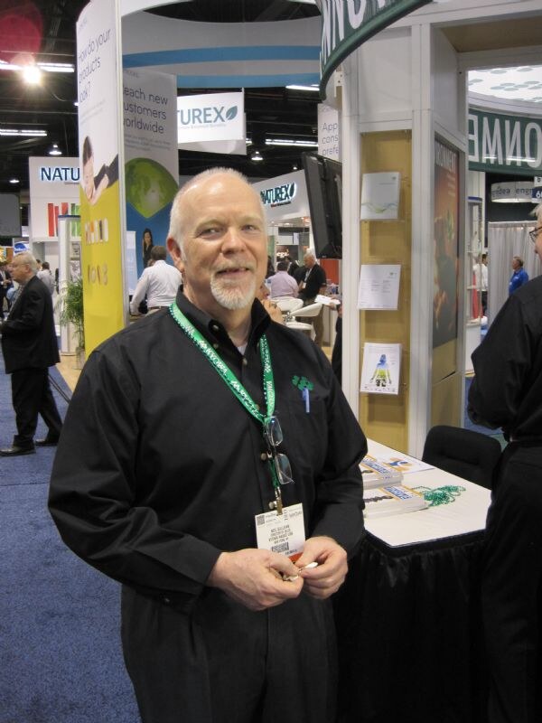 A smiling bald man with a goatee, wearing a black shirt, green lanyard and badge, stands in an exhibit aisle with Turex signs and a booth table behind him.