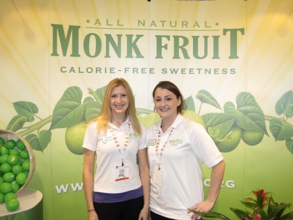 Two smiling women in white polo shirts and lanyards stand in front of a green All Natural Monk Fruit, Calorie-Free Sweetness backdrop with a bowl of green fruit.