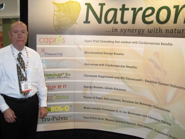 A man in glasses, white shirt and patterned tie stands beside a Natreon display listing brands including Capros, PrimaVie, ProsPlus and Tru-Fulvic.