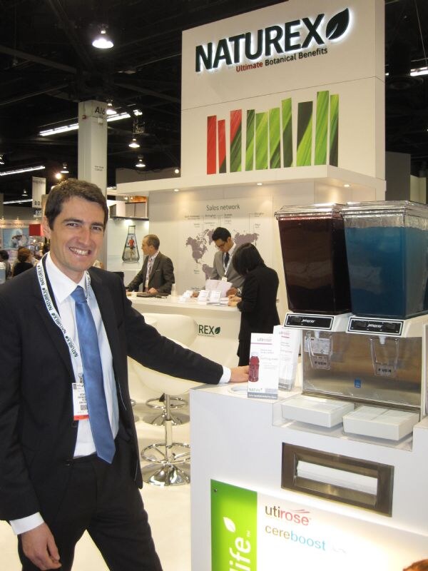 A smiling man in a dark suit and blue tie stands beside a Naturex booth with two beverage dispensers and Utirose and Cereboost signs, with people at a counter behind.