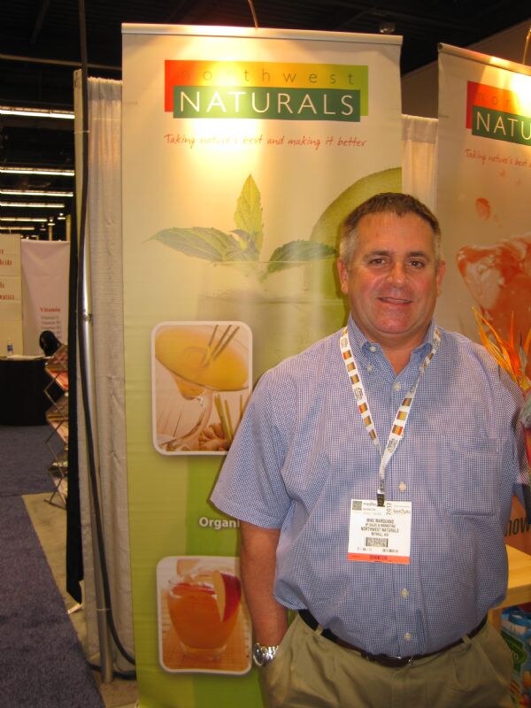 Mike Marquand of Northwest Naturals, in a blue checked shirt with a lanyard and badge, stands in front of Northwest Naturals banners showing drinks.