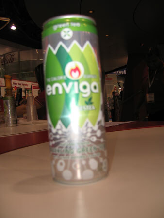 A blurry close-up of a slim Enviga green tea can in green and silver standing on a tan counter, with a blurred trade show floor and other booths in the background.