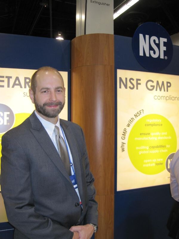 A bearded man in a gray suit and tie stands in front of an NSF booth with a round NSF logo and a lit panel reading NSF GMP compliance.