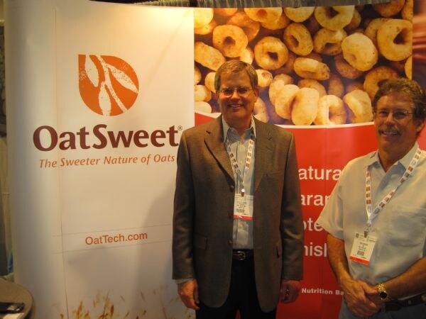 Two men with badges and lanyards stand in front of an OatSweet banner, one in a brown blazer and one in a light short-sleeved shirt, with a cereal image behind them.
