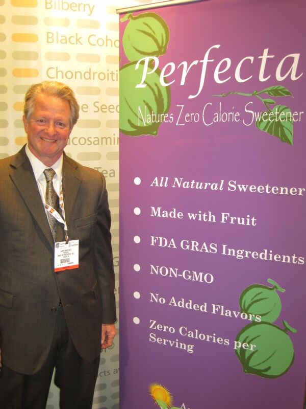 A smiling man with gray hair in a brown suit and badge stands beside a purple Perfecta banner reading Natures Zero Calorie Sweetener, with bullet points and green fruit illustrations.