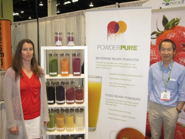 A woman in a red top and gray cardigan and a man in a blue shirt stand on either side of a PowderPure banner and a shelf of jars of colorful powders, with a strawberry graphic behind.