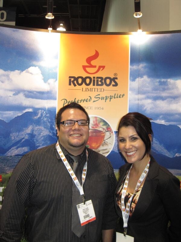 A smiling man with glasses in a pinstripe shirt and a smiling woman with dark hair in a black jacket, both wearing lanyards, stand in front of a Rooibos Limited banner with a mountain backdrop.