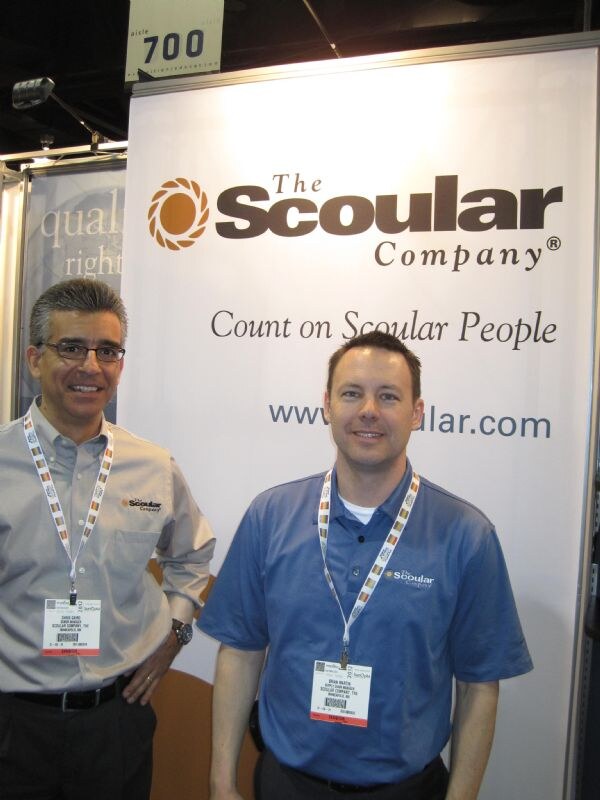 Two men wearing badges and Scoular shirts stand in front of a The Scoular Company banner reading Count on Scoular People, near a sign numbered 700.
