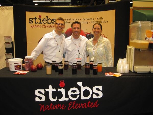 Two men and a woman in white and light shirts stand behind a black Stiebs table topped with jars of juices and concentrates and pomegranates, in front of a Stiebs Nature Elevated backdrop.