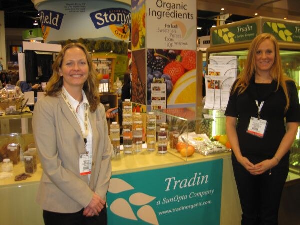 A woman in a tan blazer and a woman in black stand at a teal Tradin counter with sample jars and organic ingredients signage; a Stonyfield tub graphic appears overhead.