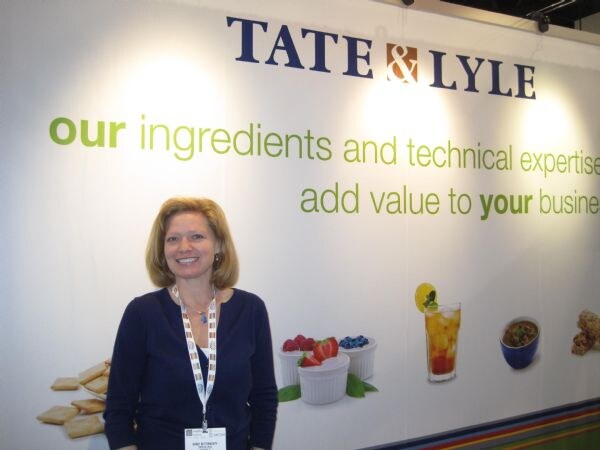 A smiling woman with shoulder-length blond hair and a lanyard stands in front of a white Tate & Lyle wall reading our ingredients and technical expertise add value to your business.