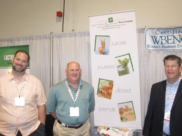 Three men wearing badges stand beside a Natural Ingredient Solutions, Vegetable Juices, Inc. banner listing juices, purees, diced and blends, with a WBENC certification sign at right.