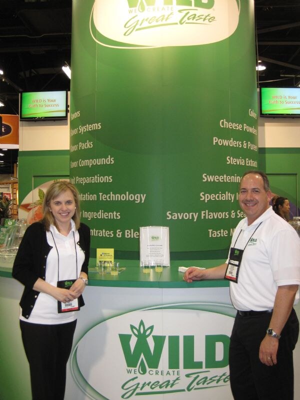 A woman in a black cardigan and a man in a white polo, both with badges, stand at a green WILD We Create Great Taste booth with screens and a list of product categories.