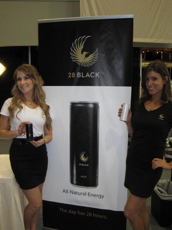 Two women, one in a white top and black skirt and one in a black dress, each hold a can beside a 28 Black banner showing an açaí can and the text All-Natural Energy.