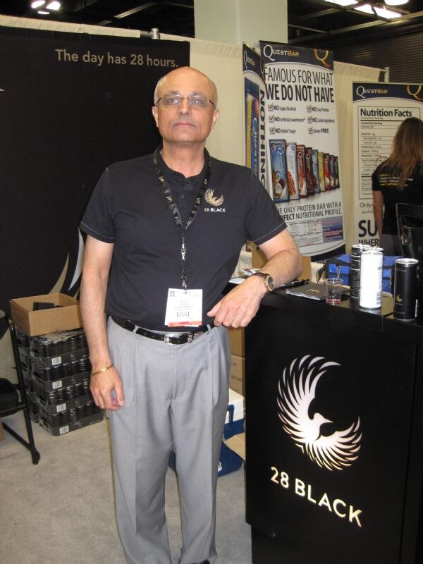 A man in a black 28 Black polo and glasses leans on a black 28 Black counter with a phoenix logo. Cases of cans sit to his left and Quest Bar banners hang behind him.