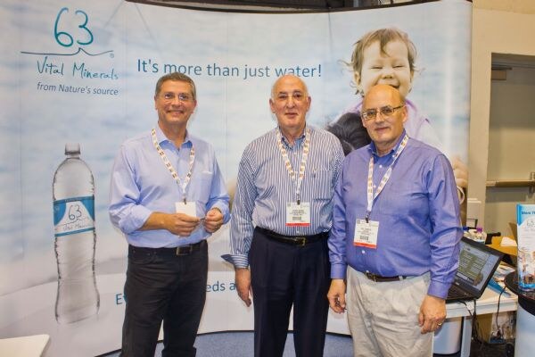 Three men in dress shirts and show badges stand side by side in front of a 63 Vital Minerals backdrop showing a water bottle and a smiling baby, with the line "It's more than just water!"