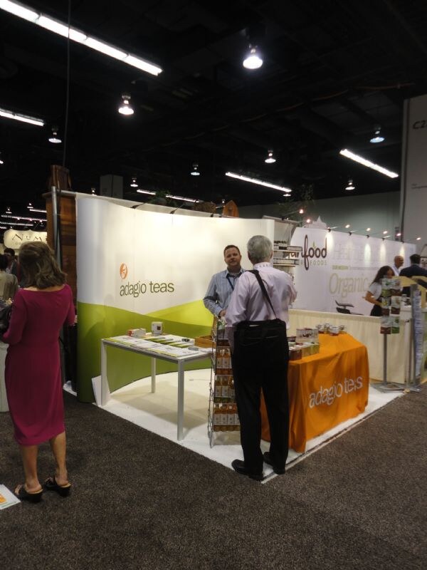 Adagio Teas booth with a curved white and green wall and an orange tablecloth. A man behind the table talks with a gray-haired man in a dress shirt; a woman in a magenta dress walks by.