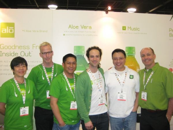 Six people in green and white alo shirts and show badges stand together in front of an alo banner reading "Aloe Vera" and "Goodness From Inside Out," with bottles of drink visible behind them.