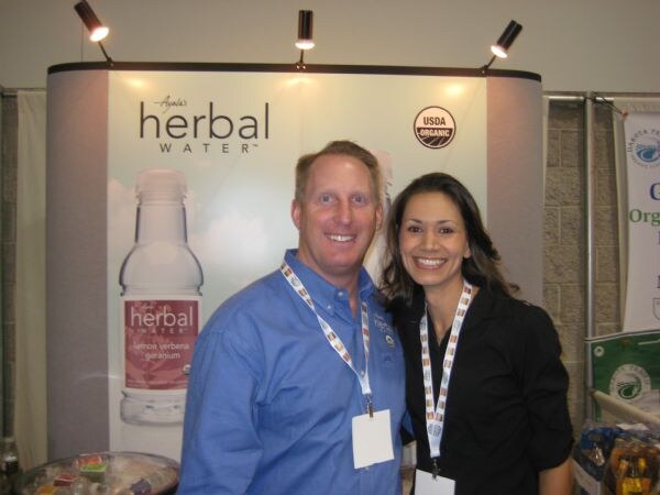 A smiling man in a blue shirt and a smiling woman in black, both wearing badge lanyards, stand in front of an Ayala's Herbal Water display with a USDA Organic seal and a bottle graphic.