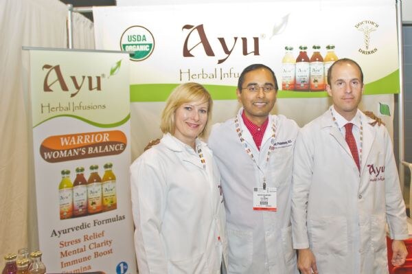 A woman and two men in white lab coats stand in the Ayu Herbal Infusions booth. A pull-up banner lists Ayurvedic formulas, and a Doctor's Drinks sign shows bottles above them.
