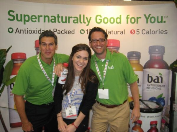 Two men in green bai polo shirts flank a woman in a gray top holding a bottle. Behind them is a banner reading "Supernaturally Good for You" and bai Antioxidant Infusions bottles.