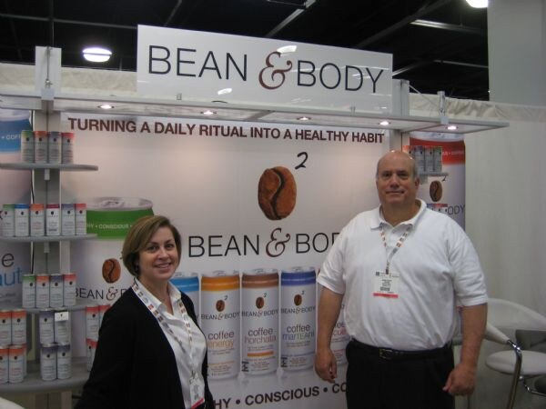 A woman in a dark cardigan and a man in a white polo stand in front of a Bean & Body display with shelves of canned coffee drinks and a banner reading "Turning a daily ritual into a healthy habit."