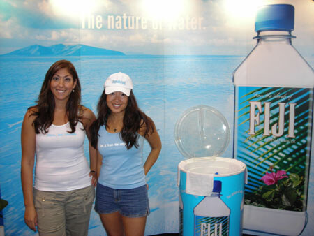 Two smiling women stand in front of a Fiji water booth with an ocean backdrop, a large Fiji bottle graphic, and a Fiji-branded cooler.