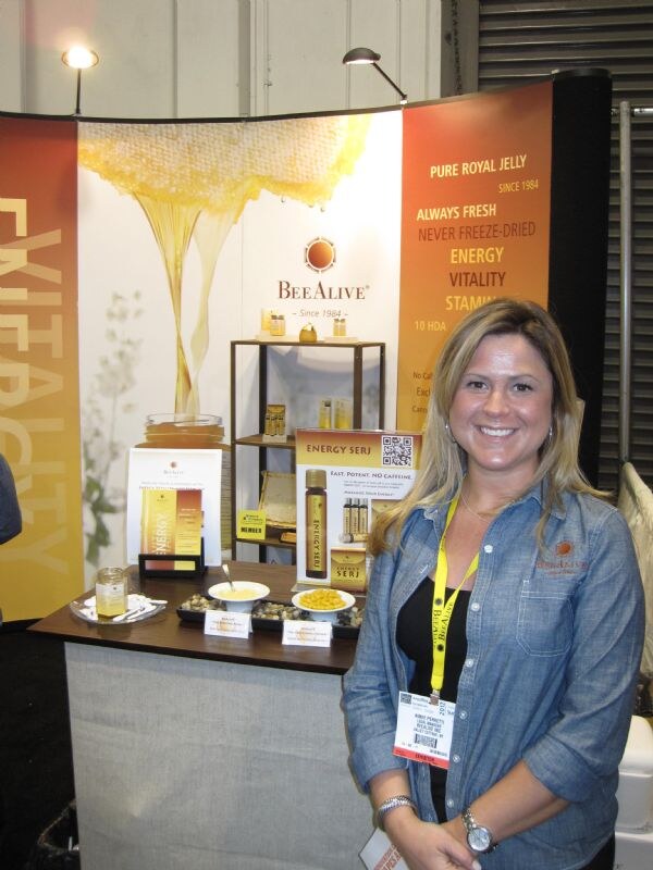 A smiling woman in a denim shirt with a yellow lanyard stands beside a BeeAlive booth with honey imagery, royal jelly signage, an Energy Serj display and small dishes on a counter.