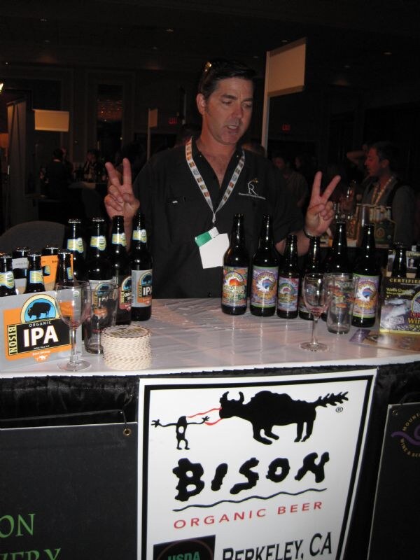 A man in a black shirt holds up two-finger peace signs behind a table of Bison Organic Beer bottles and glasses. The front sign shows a bison logo and the text Berkeley, CA.
