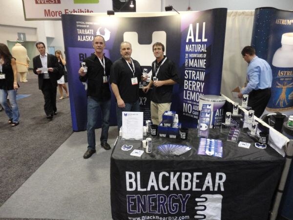 Three men with badges stand in front of a BlackBear Energy booth with a purple banner reading all natural, and a table with the BlackBear Energy logo and cans on display.