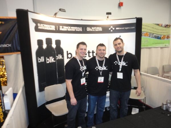 Three men in black blk. T-shirts and badges stand together in front of a blk. banner showing black bottles and the text electrolytes, antioxidants and all natural.