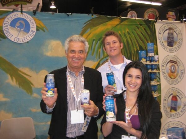 A smiling older man, a younger man and a woman hold cans and a bottle of Blue Monkey coconut water in front of a tropical backdrop and Blue Monkey Coconut Collection banners.