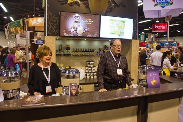 A woman and a man in dark clothing and badge lanyards stand behind a brown counter in a booth with drink dispensers, shelves of jars and screens. Blue Sky and Diamond Natural signs hang nearby.