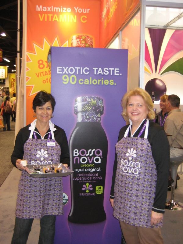 Two women in purple Bossa Nova aprons stand beside a large Bossa Nova açaí bottle display reading exotic taste, 90 calories; one holds a tray of small sample cups.
