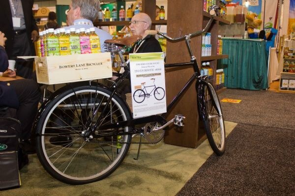 A black bicycle with a wooden Bowery Lane Bicycles crate on the back holds bottles of green and pink drinks. A sign on the bike mentions the Ito En booth. People sit behind it.