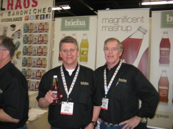 Two men in black Búcha shirts and badges stand at the Búcha Live Kombucha booth; one holds a bottle, and banners behind them read magnificent mashup.