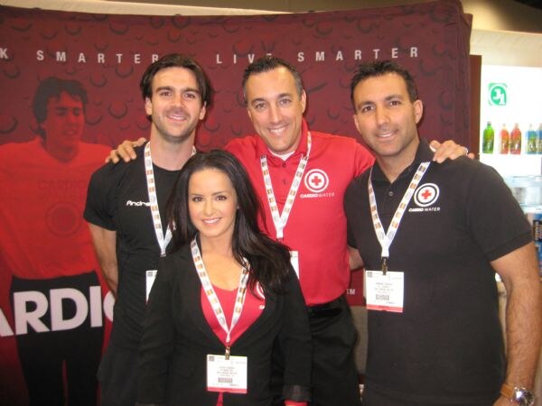Three men and a woman wearing badges pose together in front of a red Cardio Water backdrop. Two of the men wear shirts with the Cardio Water logo.