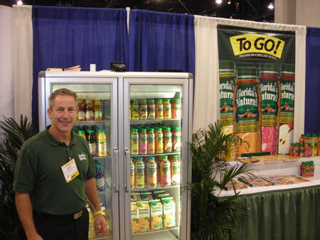 A man in a green polo shirt with a badge stands beside two glass-door coolers filled with juice bottles, next to a Florida's Natural 'To Go!' banner and a table of juice.