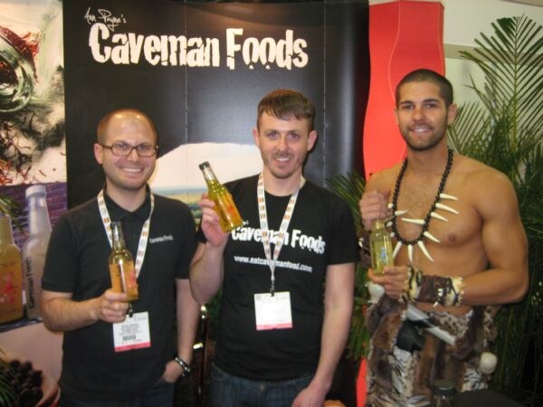 Two men in black shirts hold bottles beside a shirtless man in a fur costume and bone necklace, in front of a Caveman Foods banner.
