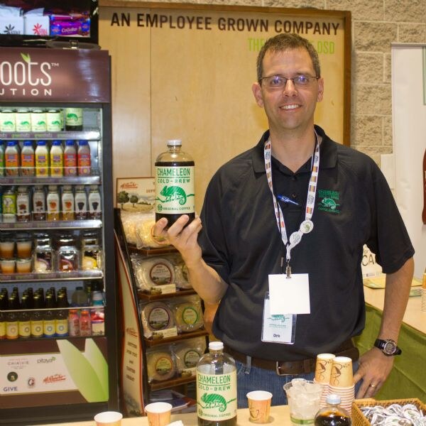 A smiling man with glasses and a name badge holds a bottle of Chameleon Cold-Brew original coffee at a booth with a refrigerated display and a sign reading an employee grown company.