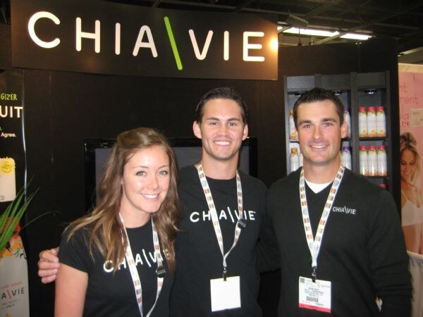 Two men and a woman in black Chia Vie shirts and lanyards smile in front of a Chia Vie sign, with bottles on shelves behind them.