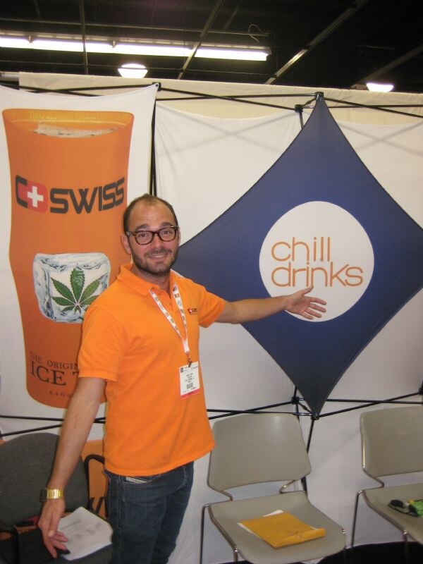 A smiling man in an orange polo and glasses gestures toward a chill drinks sign, beside a C-Swiss ice tea banner with a cannabis leaf in ice.