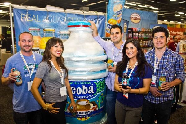 Four smiling people hold Coba bottles beside a giant Coba Horchata bottle replica, under a banner reading Real Aguas Real Sabor.