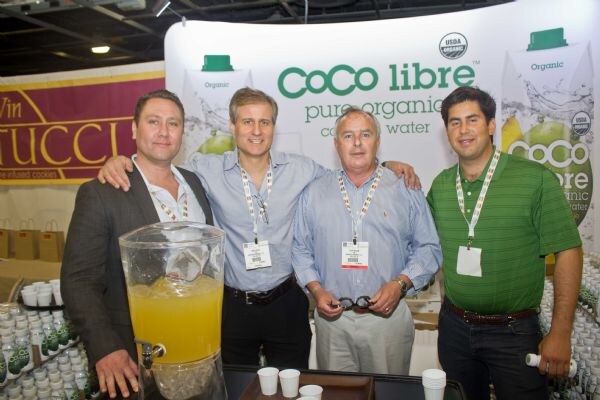 Four men stand together at a Coco Libre booth with a coconut water banner behind them and a jug of yellow drink and cups in front.