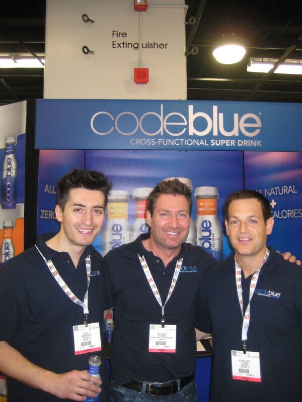 Three men in navy codeblue polos and lanyards smile in front of a codeblue sign and bottles, one man holding a bottle.