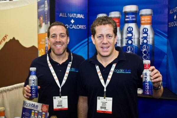 Two men in navy codeblue polos and lanyards hold codeblue bottles in front of a display of bottles reading All Natural and Zero Calories.