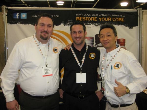 Three men wearing Core Power logo shirts and lanyards stand together in front of a banner reading Restore Your Core, one giving a thumbs up.