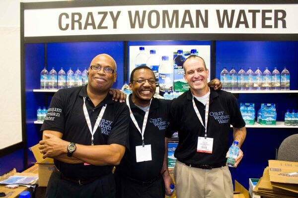 Three smiling men in black Crazy Woman Water shirts stand arm in arm in front of shelves of bottled water under a Crazy Woman Water sign.